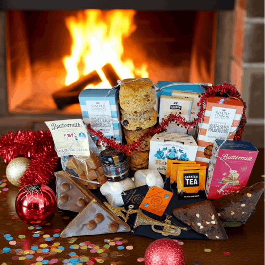 Assorted food items including scones, jam & cream, fudge, chocolate, tea bags and biscuits on a table with a fireplace in the background
