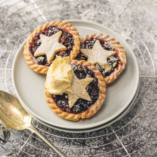 Three mince pies on a plate with a scoop of clotted cream, served on a textured surface.